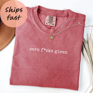 Puede incluir: Una camiseta Comfort Colors rosa polvoriento con la frase "zero f*cks given" impresa en blanco. Un collar plateado con un colgante redondo descansa sobre la camiseta. La camiseta está doblada y se muestra con un fondo neutro.