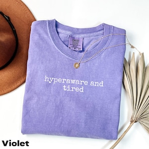 Puede incluir: Una camiseta violeta Comfort Colors con las palabras "hyperaware and tired" impresas en blanco. Un collar dorado con un colgante descansa sobre la camiseta. Un sombrero marrón y una planta seca están en el fondo.