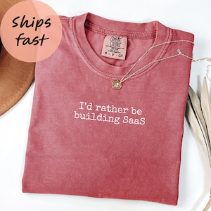 Peut inclure: Un t-shirt rose poussiéreux Comfort Colors avec l'inscription "I'd rather be building SaaS" en lettres blanches. Un collier argenté avec un pendentif circulaire repose sur le t-shirt. Le t-shirt est plié et porte un autocollant rond indiquant "Expédié rapidement".