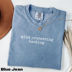Peut inclure: Un t-shirt bleu jean avec l'inscription blanche « mind processing backlog ». Un collier doré avec un pendentif repose sur le t-shirt. Le t-shirt est plié et présenté avec un chapeau marron.