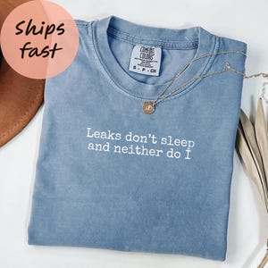 May include: A blue Comfort Colors t-shirt with the text "Leaks don't sleep and neither do I" in white. A gold necklace with a pendant rests on the shirt. The shirt is folded on a surface.