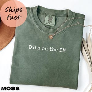 May include: Moss green t-shirt with the text "Dibs on the DM" in white. A gold necklace with a pendant is draped across the shirt. The shirt is folded on a white surface. The word "MOSS" is printed in white at the bottom.