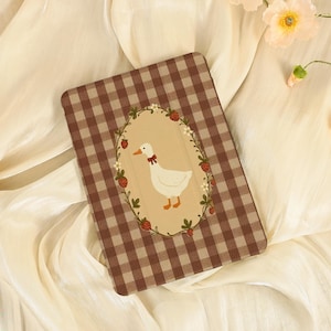 May include: A rectangular notebook cover with a brown and white gingham pattern. The central oval design depicts a white goose wearing a red bow tie, framed by a wreath of strawberries and flowers. The background is a cream-coloured fabric.