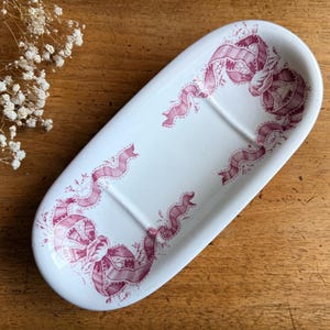 Peut inclure: Un porte-savon ovale en céramique blanche orné d'un motif décoratif bordeaux de rubans et de nœuds. L'objet présente une finition lisse et brillante, un centre légèrement creusé et deux petites nervures en relief sur la surface centrale.