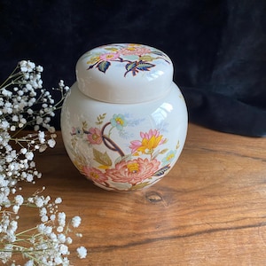 Könnte beinhalten: Weiße Keramikdose mit einem Blumendesign mit rosa, gelben und blauen Blumen. Der Deckel ist ebenfalls mit einem Blumendesign verziert.