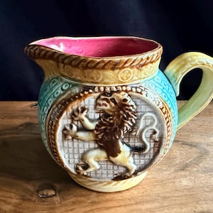 Peut inclure: Pichet en céramique vintage avec un emblème de lion. Le pichet présente un intérieur rose, une anse jaune et un corps turquoise avec des détails en relief. Le lion est brun et beige, sur fond blanc.