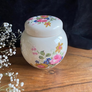 May include: A white ceramic jar with a floral design. The jar has a lid and is decorated with pink, yellow, and blue flowers.