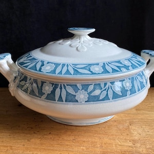 May include: A white and blue ceramic serving dish with a lid. The dish has a floral and leaf pattern in blue. The handles and lid finial are white. The dish is on a wooden surface.