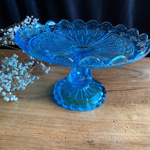 Può includere: Un porta torta in vetro blu con un bordo smerlato e un design intricato. Il supporto ha una base a piedistallo ed è perfetto per esporre dessert o altre leccornie.