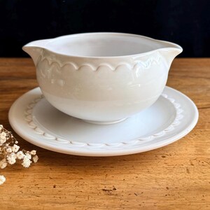 May include: Cream-coloured gravy boat and matching saucer. The boat has a scalloped rim and a spout. The saucer also features a scalloped edge. A classic serving piece for sauces and gravies, perfect for a dinner table.