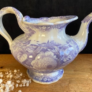 Puede incluir: Jarra de cerámica vintage con un diseño floral en tonos morados y blancos. La jarra tiene un asa curva y un pico con forma de pájaro decorativo. La base es festoneada y el diseño es ornamentado.