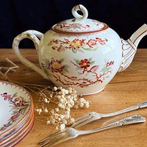 May include: A white porcelain teapot with a lid, decorated with red, green, and yellow floral and bird motifs. A stack of matching plates and two silver-toned metal forks are positioned nearby, accompanied by a small sprig of dried white flowers.