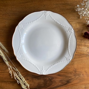 May include: A white ceramic plate with a gold rim and embossed floral design. The plate has a scalloped edge and is set on a wooden surface. Decorative elements include dried flowers and wheat stalks.