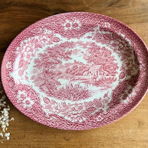 May include: An oval ceramic platter featuring a pink and white transferware pattern. The center depicts a rural landscape with a stone bridge, a house, and trees. The wide rim is decorated with a detailed floral and berry border design.
