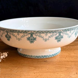 May include: A white ceramic pedestal bowl featuring a teal green floral garland pattern around the exterior rim and base. The bowl has a smooth, rounded shape with a wide opening and a sturdy circular footed base, showcasing a classic design.