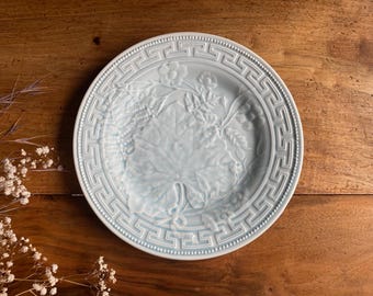 May include: A light blue ceramic plate with a detailed embossed floral and leaf design. The plate features a Greek key border and is set against a warm wooden surface. Decorative tableware.