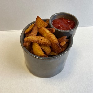 May include: A gray ceramic bowl with a smaller bowl for dipping sauce. The bowl is filled with crispy potato wedges and the smaller bowl contains red ketchup.