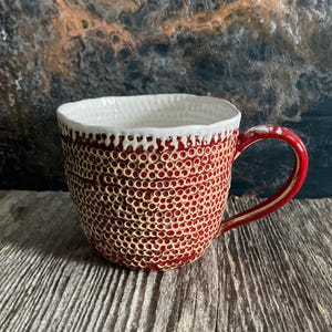 May include: A ceramic mug with a textured, red exterior and a white interior. The mug has a unique pattern of small, evenly spaced holes, and a red handle. The rim is white with a textured surface. The mug is designed for hot drinks.