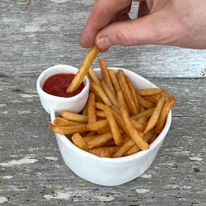 May include: A white ceramic bowl filled with golden-brown chips, with a small attached dish of red ketchup. A hand dips a chip into the ketchup. The bowl sits on a weathered wooden surface.