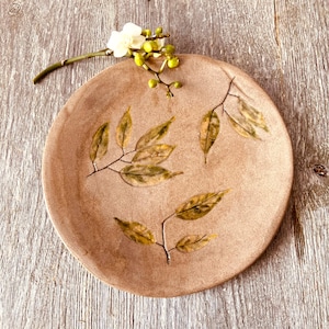 Peut inclure: Une assiette ronde en céramique de couleur beige avec un motif de feuilles. Les feuilles sont peintes dans des tons de vert et de jaune. Une petite branche de fleurs blanches et de boutons verts repose sur le bord de l'assiette.