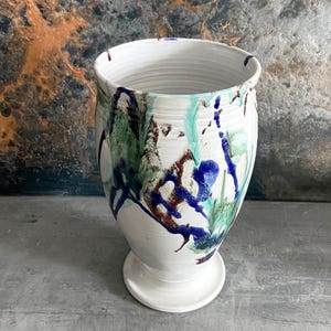 May include: White ceramic vase with a wide opening and a pedestal base. The vase is adorned with dripped patterns in shades of blue, green, and brown. The interior is white, and the exterior has a textured finish.