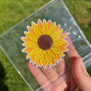 May include: Embroidered sunflower patch with yellow petals and a brown centre, set against a white border. The patch is held in a clear plastic sleeve, with a green grassy background.