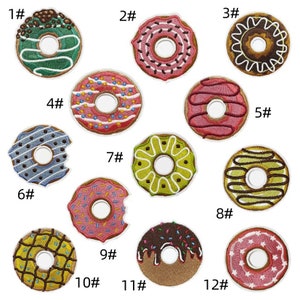 May include: A selection of twelve embroidered donut patches. Each patch features a different design, with various colours and toppings, including sprinkles and chocolate. The patches are numbered 1 to 12. The patches are suitable for clothing.