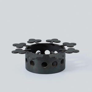 May include: A dark grey metal candle holder with a circular base and small, round holes. The top edge has decorative clover-shaped cutouts. The candle holder is designed to hold a small candle.