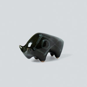 May include: A dark green ceramic figurine of a rhinoceros with a glossy finish. The body features a light green, textured geometric pattern consisting of a central circle enclosed within a diamond shape, with additional lines extending across the back.