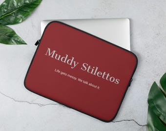 May include: A dark red laptop sleeve with black trim featuring the white text Muddy Stilettos and the smaller white text Life gets messy. We talk about it. The sleeve is partially open, revealing a silver laptop inside the protective case.