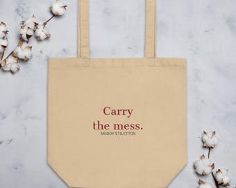May include: A beige canvas tote bag with two shoulder straps. The front features the text Carry the mess. in dark red, with the words MUDDY STILETTOS printed in smaller dark gray letters directly underneath the main phrase.