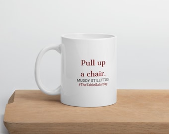 May include: A white ceramic mug with a handle on the left side. The text printed on the front reads: Pull up a chair. MUDDY STILETTOS #TheTableSaturday. The text is in dark red and grey lettering against the plain white surface of the mug.