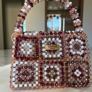 Peut inclure: Un sac à main perlé avec une poignée. Le sac est fait de petites perles colorées dans les tons de blanc, rose et bordeaux. Le sac a une forme rectangulaire avec une étiquette dorée qui dit "KASKO BAGS".