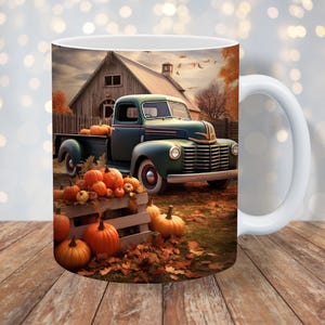 May include: White ceramic mug featuring a fall scene with a vintage teal pickup truck loaded with pumpkins. A wooden crate overflowing with pumpkins sits in front of the truck. The background shows a barn and autumn foliage.