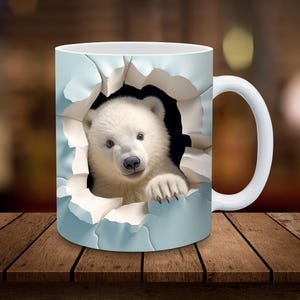Könnte beinhalten: Weiße Keramiktasse mit einem 3D-Bild eines Eisbärenjungen, das aus einem Loch in der Tasse schaut. Das Bärenjunges hat ein weißes Fell und schwarze Augen und Nase.