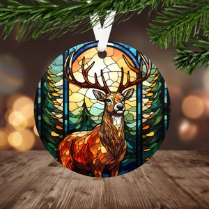 May include: A stained glass style Christmas ornament featuring a deer standing in a forest setting. The deer is brown with a large rack of antlers. The background is a colorful stained glass design with shades of blue, green, yellow, and orange.