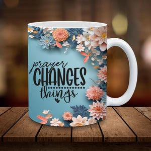K&ouml;nnte beinhalten: Wei&szlig;e Keramiktasse mit blauem Hintergrund und floralem Design. Der Text "prayer CHANGES things" ist in Schwarz auf die Tasse gedruckt.