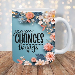 K&ouml;nnte beinhalten: Wei&szlig;e Keramiktasse mit blauem Hintergrund und Blumenmuster. Der Text "prayer CHANGES things" ist in Schwarz auf die Tasse gedruckt.