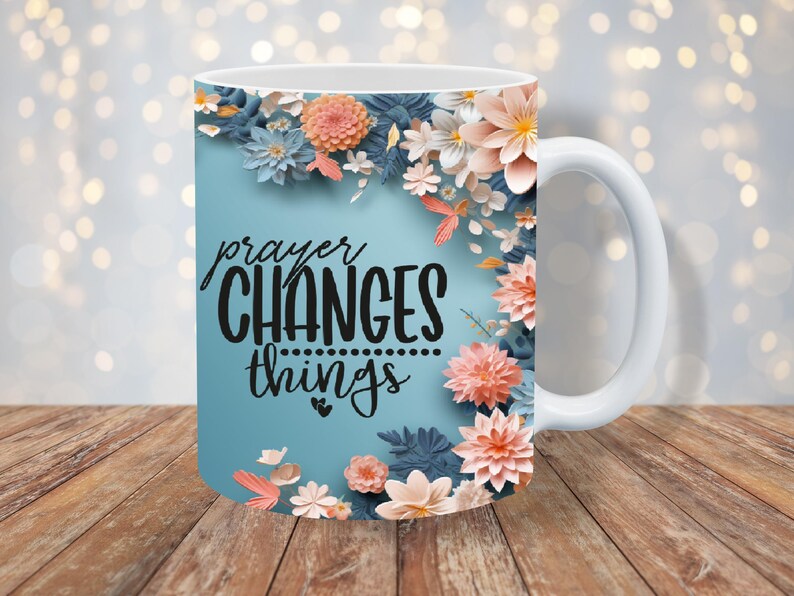 K&ouml;nnte beinhalten: Wei&szlig;e Keramiktasse mit blauem Hintergrund und Blumenmuster. Der Text "prayer CHANGES things" ist in Schwarz auf die Tasse gedruckt.