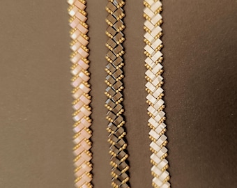 Puede incluir: Tres pulseras de cuentas en fila. La primera pulsera es de color rosa claro con cuentas doradas. La segunda pulsera es negra con cuentas doradas. La tercera pulsera es blanca con cuentas doradas. Las pulseras están sobre una superficie marrón oscuro.