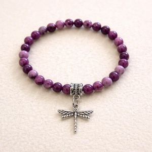 May include: A purple beaded bracelet with a silver dragonfly charm. The bracelet features round, purple beads and a decorative silver bead. The dragonfly charm is detailed with wings and a slender body. The bracelet is a piece of jewelry.