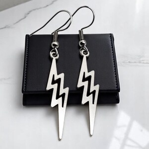 May include: Silver-colored lightning bolt earrings with a hook closure. The earrings are displayed on a black, rectangular object, set against a white and gray marble surface. The earrings have a modern, edgy design.