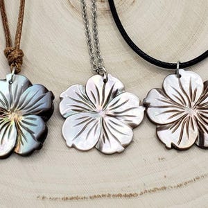 May include: Three necklaces with flower pendants. The pendants are made of mother of pearl and are carved in the shape of a flower. The necklaces are on a wooden surface.