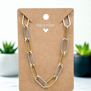 May include: A handmade necklace with a chain link design. The necklace features alternating gold and silver-coloured links. The necklace is displayed on a brown card with the word "Handmade" and a heart symbol.