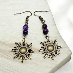 Puede incluir: Pendientes de sol morado y dorado con un dije de sol colgante y dos cuentas moradas.