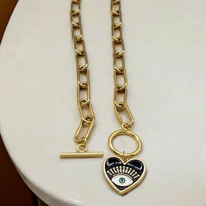 May include: Gold-toned necklace with a heart-shaped pendant featuring an eye design. The pendant is black with a gold border and a central green eye. The necklace has a toggle clasp and a chain link design.