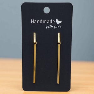 Puede incluir: Un par de pendientes dorados en forma de barra, presentados en una tarjeta negra. La tarjeta tiene el texto "Handmade with love" en blanco. Los pendientes son largos y finos.