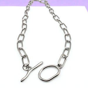 May include: Silver-toned chain necklace with an oval-shaped clasp. The necklace features a series of elongated, rectangular links. The clasp is a bar and loop design. The jewelry is displayed on a white surface with a purple background.