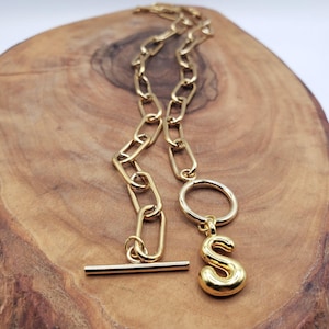 May include: A gold-coloured necklace with a large link chain and a toggle clasp. The necklace features a pendant with the letter "S" and a circular ring. The jewellery is displayed on a wooden surface.