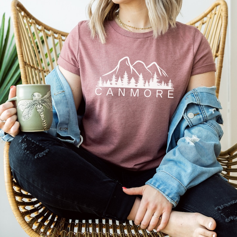 May include: A woman wearing a mauve t-shirt with a white mountain and forest graphic and the text "Canmore". She is wearing a denim jacket and black jeans.